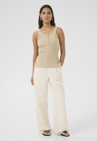 Beige räfflad tanktop med rund halsringning och knappdetalj, i kombination med löst sittande krämfärgade byxor och svarta flip-flop sandaler.