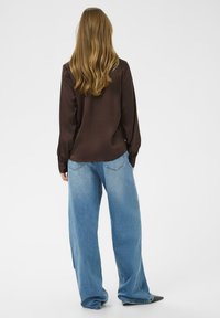 Bluză lungă din satin maro cu mâneci lungi, asortată cu blugi din denim ușori, de mari dimensiuni, având o croială relaxată și o ușoară decolorare. Pantofi negri cu vârf ascuțit.
