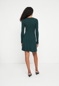 Femme aux cheveux bouclés portant une robe à manches longues ribbed verte foncée et des sandales à talons noires, se tenant de dos contre un fond blanc.