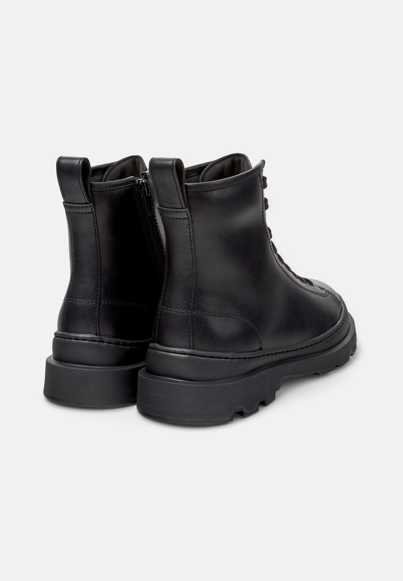 Een paar zwarte leren enkellaarsjes met dikke rubberen zolen, achterste treklussen, achterste ritsen en vetersluiting op een witte achtergrond.