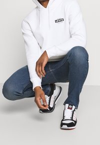 Person som bär en vit Levi's hoodie, blå jeans och svart-vita Adidas sneakers, knäböjer med en hand som justerar en skosnöre.