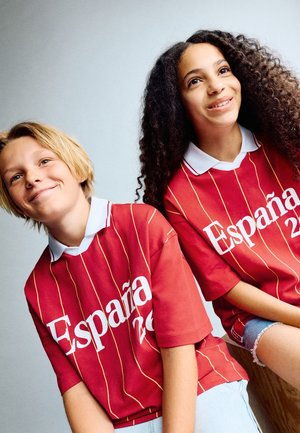 Twee glimlachende tieners met rode España 2022 sportshirts, zittend tegen een effen lichte achtergrond.