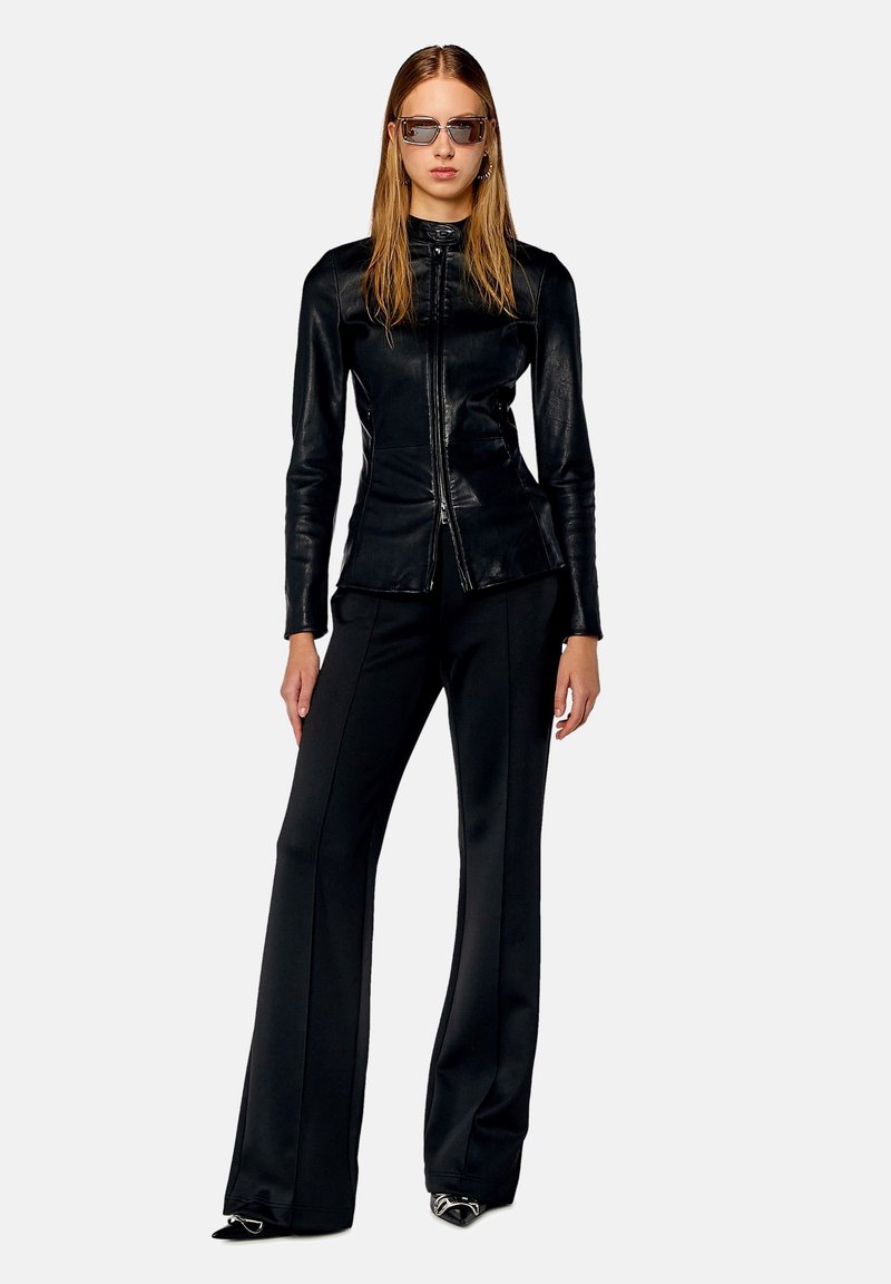 Frau in schwarzer, Leder-Jacke mit Reißverschluss, schwarzen weiten Hosen, spitzen Schuhen und rechteckiger Sonnenbrille, die vor einem einfarbigen Hintergrund steht.