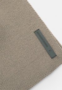 Knitted beige fabric with a textured pattern, featuring a black rectangular label embossed with "ALTERNATIVE CULTURE" on one edge.