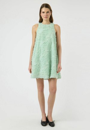 Vrouw staand, gekleed in een mouwloze, muntgroene, getextureerde A-lijnjurk en zwarte balletschoenen, tegen een effen witte achtergrond.