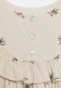 Jasnobeżowa tkanina z delikatnymi teksturowanymi kropkami, mały nadruk kwiatowy, trzy iryzujące guziki oraz marszczenie z falbanką na dekolcie odzieży.