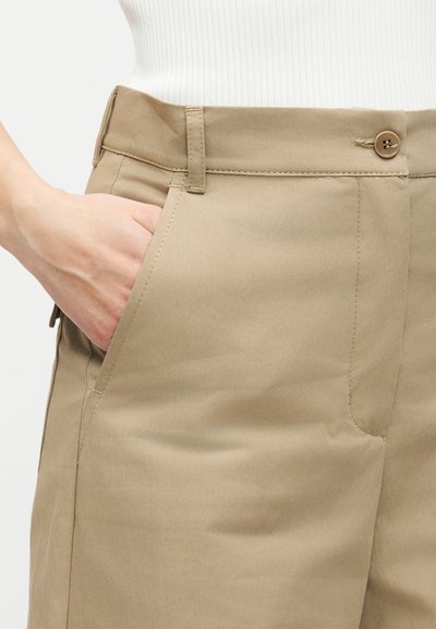 Χέρι στην μπροστινή τσέπη μπεζ προσαρμοσμένου παντελονιού που φοριέται με λευκό ριμπ μπλουζάκι, εμφανίζοντας κουμπί και θηλιές για ζώνη στο παντελόνι.