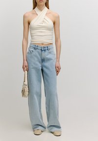 Crème haltertop met gedraaide ontwerp, gecombineerd met lichtblauwe wijde jeans. Model houdt een kleine beige tas vast en draagt beige schoenen.
