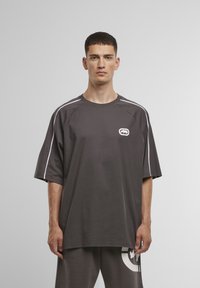 Overdimensioneret mørkegrå t-shirt med hvid kontrastsyning og et logo på brystet, lavet af bomuld, med en afslappet pasform og korte ærmer.