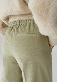 Hellgrüne Cordhosen aus einem strukturierten Stoff, mit einem elastischen Bund und einem kleinen Rückentaschendetail, teilweise verdeckt von einem beigefarbenen, flauschigen Oberteil.