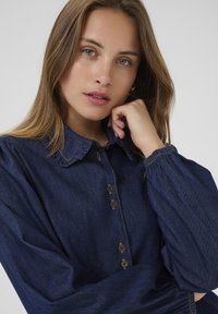 Dark denim blouse featuring a ruffled collar, button-down front with metallic buttons, and billowy sleeves with elastic cuffs.