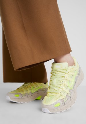 Personne portant un pantalon large marron et des baskets jaunes Nike avec des accents vert néon, debout sur une surface blanche.