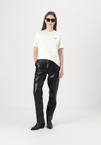Vit kortärmad t-shirt med en logo, kombinerad med glänsande svarta konstläderbyxor och svarta klackade ankelskor. Modellen har på sig solglasögon.