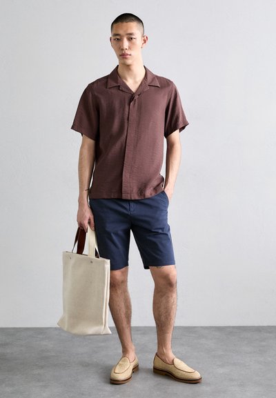 Braunes kurzärmliges Hemd, marineblaue Shorts, Strohschnürschuhe und eine cremefarbene Tragetasche mit Stoffgriffen, stehend auf einem grauen Boden.