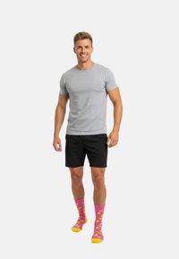 Smilende mand står iført en lys grå t-shirt, sorte shorts og lysende pink sokker med gule bananmønstre på en hvid baggrund.