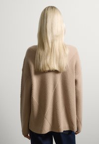 Maglione a costine di un beige chiaro con una vestibilità ampia, caratterizzato da un motivo a diamante e maniche larghe, indossato sopra jeans blu scuro.