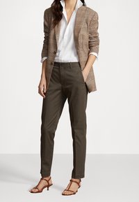 Kvinna i brun rutig blazer, vit skjorta med knappar, olivgröna byxor och bruna klacksandaler, står med ena handen i fickan.