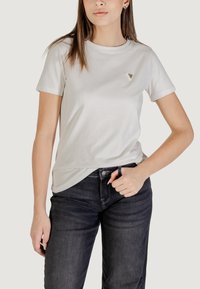 T-shirt grigio chiaro a maniche corte con scollatura rotonda e un piccolo logo triangolare sul petto, abbinato a jeans blu scuro.