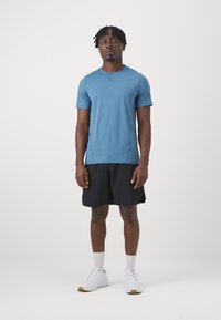 Μπλε αθλητικό μπλουζάκι με κοντό μανίκι συνδυασμένο με μαύρα σορτς. Η μπλούζα είναι λείος και το σύνολο διαθέτει λευκά παπούτσια με ανοιχτό καφέ σόλες.