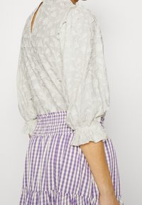 Blouse blanche brodée avec manches bouffantes et poignets élastiques, associée à une jupe à volants rayée violet et blanc.