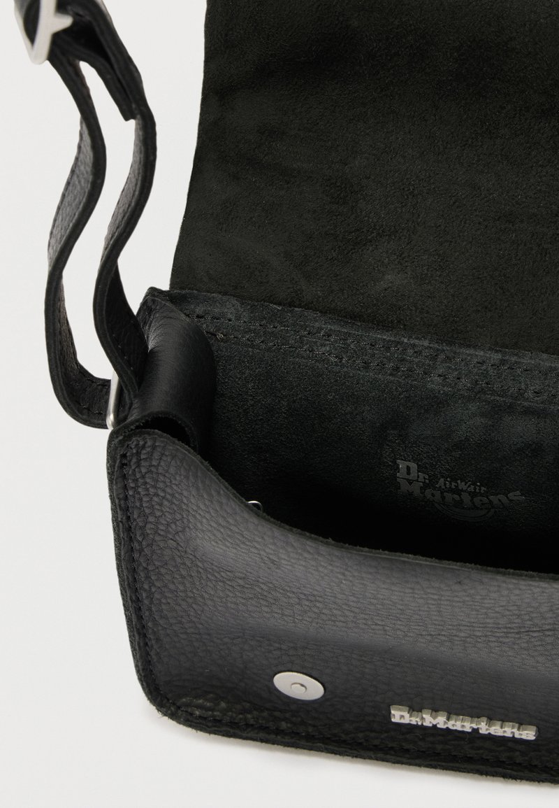 Schwarze Leder-Umhängetasche mit geöffnetem Klappdeckel, die ein Wildlederinnere und einen silbernen Magnetverschluss sowie das Dr. Martens Logo auf der Vorderseite zeigt.
