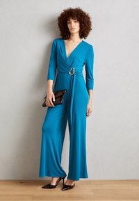 Teal jumpsuit med dyp V-hals, trekvartlange ermer, og et stort gulltonet belte i midjen. Båret med svart clutch og høye hæler.