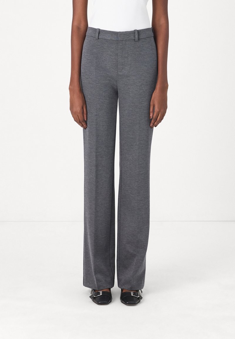Femme portant un pantalon gris droit et des chaussures noires à bouts carrés, debout devant un fond blanc uni.