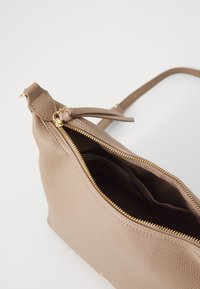 Bolso de cuero color marrón claro con una superficie texturizada, con un zipper dorado y un detalle anudado en la correa. El interior está forrado y es espacioso.