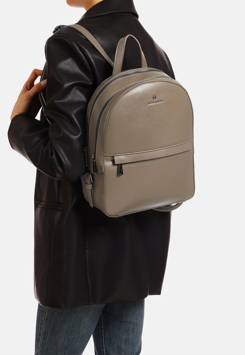 Szara skórzana plecak o zaokrąglonym kształcie, z przednią kieszenią na zamek i prostymi detalami, noszony na ciemnej kurtce.