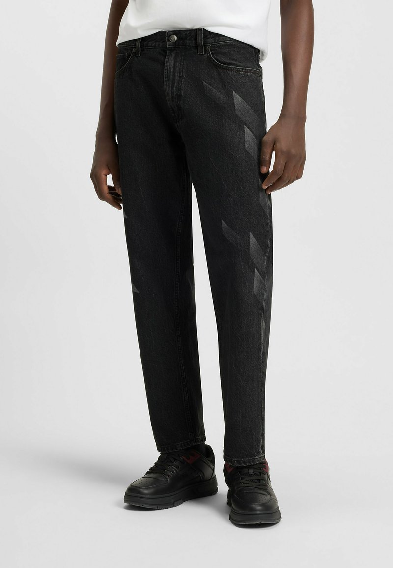 Black denim jeans with a straight-leg design, featuring diagonal stripes in a lighter shade. Worn with black sneakers.