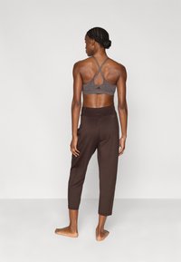 Αθλητικό σουτιέν σε σκούρο γκρι με λουράκια που διασταυρώνονται και λεπτομέρεια με άνοιγμα στην πλάτη, συνδυασμένο με καφέ παντελόνια cropped και γυμνά πόδια.