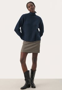 Donkerblauwe gebreide trui met hoge hals, geribde zoom en mouwen, gecombineerd met een bruin en crèmekleurig geruit minirok en zwarte kniehoge laarzen.