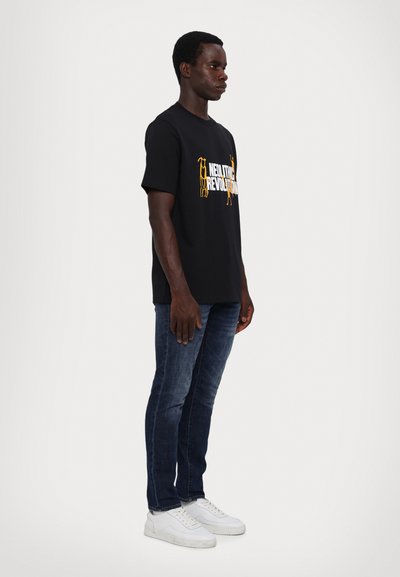 Sort bomulds T-shirt med hvid og gul grafisk tekst, parret med mørkeblå slim-fit jeans og hvide sneakers, set fra siden.
