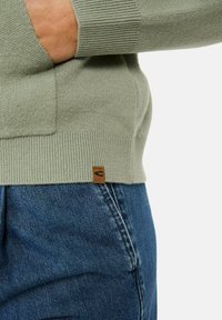 Jasnozielony sweter dzianinowy z prążkowanymi mankietami i dołem, z małą brązową skórzaną metką. W połączeniu z niebieskimi dżinsami.