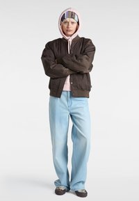 Braune gesteppte Bomberjacke, pinke Kapuzenjacke darunter, hellblaue weite Jeans, gemusterter Haarband und dunkle Schuhe mit strukturierten Akzenten.