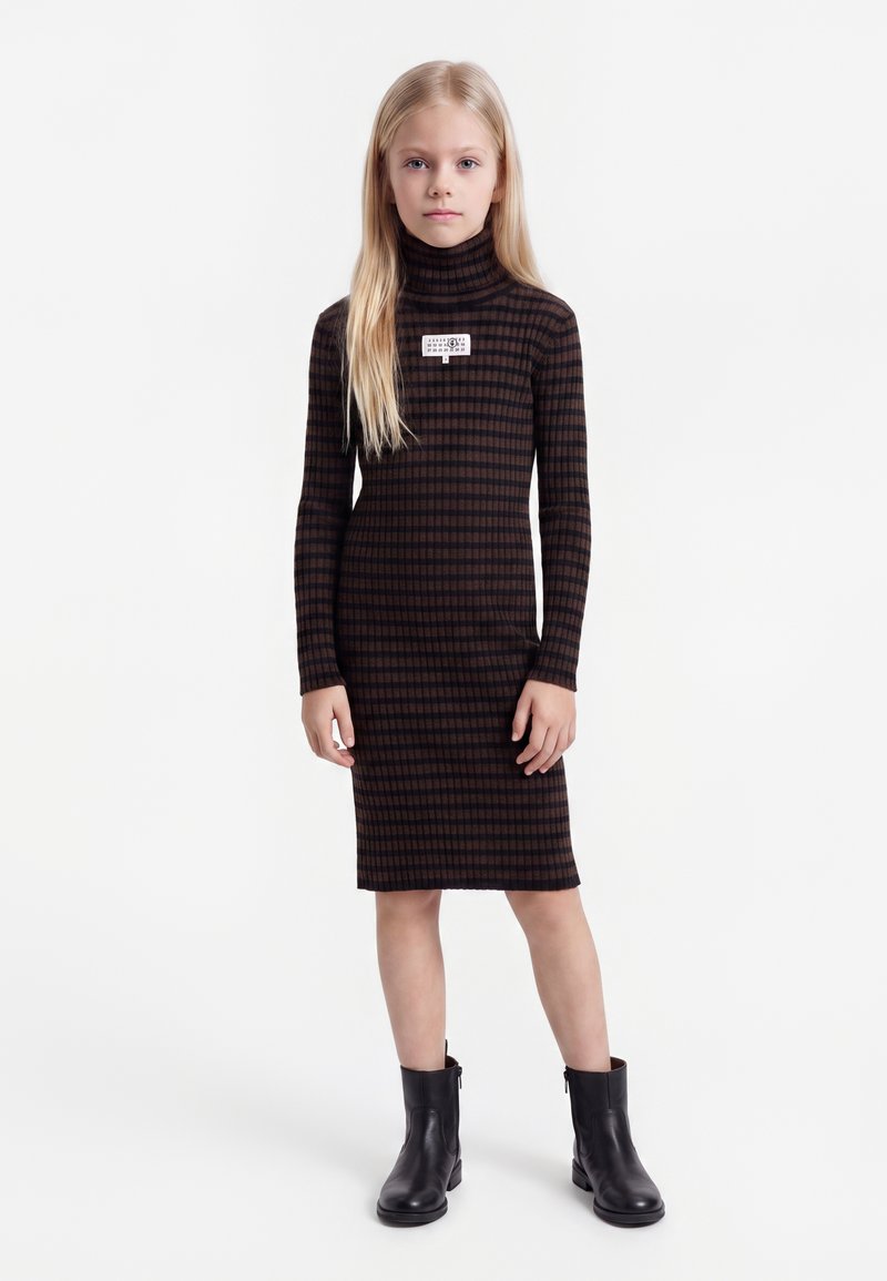 Jeune fille aux longs cheveux blonds porte une robe à col roulé à carreaux marron et noir et des bottines noires, debout sur un fond blanc.