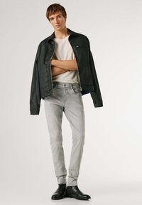 Mladý muž v černé kožené bundě přes béžové tričko, světle šedé úzké džíny a černé kožené boty, stojící se založenýma rukama.