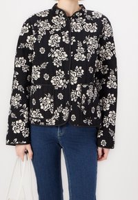Czarna pikowana kurtka z białym kwiatowym wzorem, z kołnierzem mandaryńskim, dwoma przednimi kieszeniami i zapięciem na guziki.