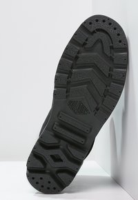 Semelle en caoutchouc noir avec un motif en zigzag texturé, comportant des découpes circulaires et rectangulaires pour une meilleure traction et flexibilité. Logo de la marque visible.