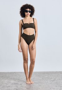 Svart bikinitopp med prikker, brede stropper, bandeau-top og høyt liv. Modell iført svarte rektangulære solbriller, står på en teksturert overflate.