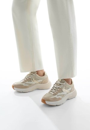Beige mesh and suede sneakers with white soles, featuring a textured design and gold accents. Worn with loose white pants.
