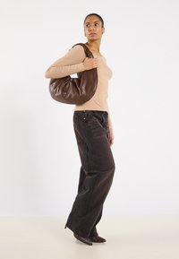 Borsa a tracolla in pelle marrone con un design morbido e arrotondato. Indossata con una maglia beige aderente a coste e jeans scuri a gamba larga.