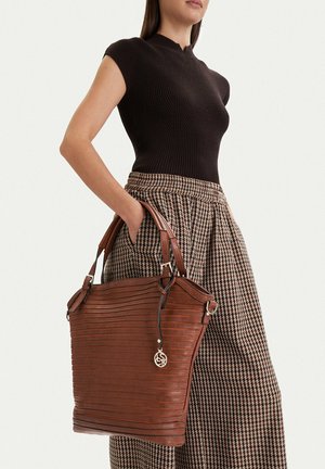 Žena v černém tílku a károvaných kalhotách drží velkou hnědou koženou kabelku s dvojitými úchyty a kulatým zlatým přívěskem.