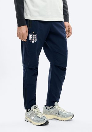 Personne portant un pantalon d'entraînement de football England bleu marine avec logo et des chaussures de course Nike argentées sur fond blanc.
