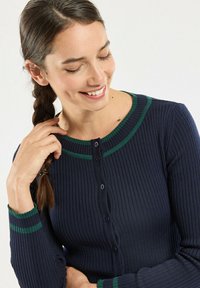Cardigan côtelé bleu marine avec accents à rayures vertes, doté d'un col rond, d'une fermeture à boutons et de manches longues. Texture douce et coupe ajustée.