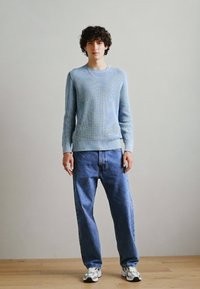 Helles blaues strukturiertes Strickpullover mit Rippbündchen, kombiniert mit locker sitzenden blauen Denim-Jeans. Das Modell trägt weiße Sneaker und silberne Accessoires.