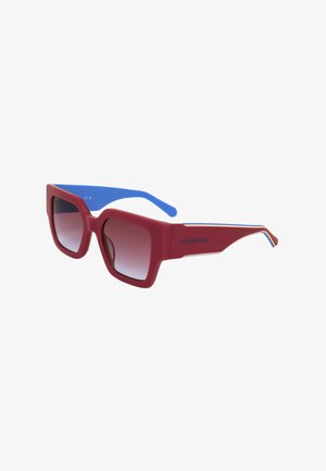 Occhiali da sole con montatura quadrata rossa, aste interne blu e lenti scure sfumate, logo Calvin Klein Jeans sul lato.