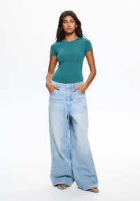 Zelený přiléhavý top s krátkým rukávem, který je zkombinován s oversized světle modrými džíny z denimu s širokými nohavicemi a viditelnými stehovými detaily.