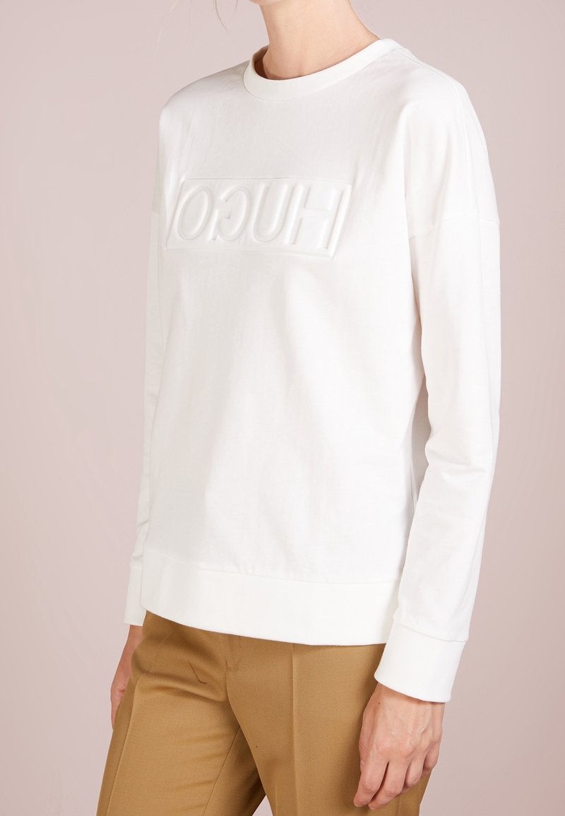 Personne portant un sweat-shirt blanc à manches longues avec un texte en relief et un pantalon beige, vue des épaules aux hanches sur un fond neutre.