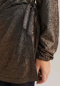 Mirdzošs, tumšs metālisks blūze ar garām piedurknēm, ar teksturētu virsmu un piesietu vidukli. Gluda auduma ar smalku spīdumu un riekšķu locījumu.